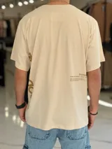 Remera oversize color beige, con mangas cortas y cuello redondo. Presenta un diseño estampado con texto en la parte trasera y lateral, de corte holgado y estilo urbano.