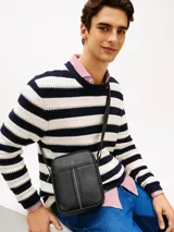 Morral de cuero sintético con acabado granulado color negro, de Tommy Hilfiger. Cuenta con un compartimento principal con cierre de cremallera, bolsillo frontal decorado con una franja tricolor vertical, correa cruzada ajustable y el logo metálico de la marca en la parte superior.