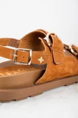 Sandalia de gamuza color tostado con base de goma marrón, diseño de tiras anchas con hebillas metálicas doradas y ajuste en el talón.