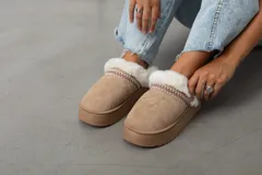 Pantuflas tipo mule con plataforma, confeccionadas en gamuza color beige. Presentan un borde superior de piel sintética blanca con detalle de cinta tejida decorativa y suela de goma a tono.