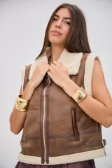 Chaleco de estilo aviador en color camel, confeccionado en material efecto cuero con interior y bordes de corderito sintético color crema. Presenta cierre frontal, cuello solapa y detalles de hebillas metálicas en los laterales y el cuello.