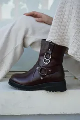 Bota de caña media confeccionada en cuero color marrón oscuro, con suela track negra de goma. Presenta un diseño estilo western con correas decorativas, remaches metálicos y argollas plateadas en el lateral.
