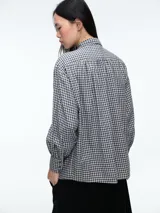 Camisa de corte clásico con estampado de cuadros vichy en tonos azul y blanco. Presenta cuello camisero, cierre frontal con botones ocultos y mangas largas con puños abotonados. Incluye un detalle de broche metálico dorado en el cuello.