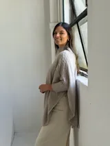 Poncho de tejido de punto color visón, con cuello alto y diseño de trenzas verticales. Presenta mangas cortas ajustadas y un corte holgado y redondeado en el ruedo.
