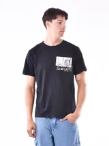 Remera negra de manga corta y cuello redondo, con un pequeño estampado rectangular en el pecho que simula una fotografía en blanco y negro de una ciudad con texto manuscrito debajo.