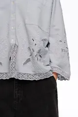 Camisa de manga larga color celeste, de corte holgado, con cuello clásico, cierre frontal de botones, bolsillo en el pecho y detalles de bordados florales y encaje en el ruedo y los puños.
