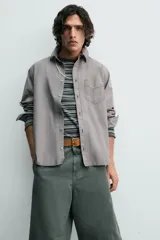 Camisa blanca de algodón con corte relajado, cuello solapa, manga larga con puño abotonado y bolsillo de plastrón en el pecho. Presenta bajo asimétrico con aberturas laterales y cierre frontal de botonadura.