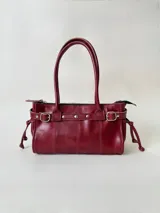 Cartera de cuero de tamaño medio con estructura rígida. Presenta una tira decorativa con tachas y hebillas metálicas en los extremos, además de lazos laterales. Cuenta con doble asa para llevar al hombro y cierre superior.