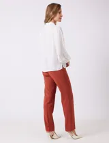 Camisa de gasa blanca con cuello solapa y cierre frontal de botones. Presenta un delicado galón de encaje festoneado a lo largo de la cartera frontal. Sus mangas largas son ligeramente abullonadas, con insertos de puntilla horizontal y puños elásticos fruncidos.