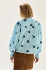 Blusa de algodón en color celeste con estampado floral en negro. Presenta cuello alto con volados y borde en color rojo, mangas abullonadas con puños elásticos tipo smock y cierre frontal con botones.