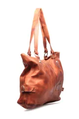 Cartera tipo shopper de cuero color marrón tostado, con doble asa de hombro, costuras decorativas centrales, detalles de tiras laterales con hebilla y un parche de cuero con logo grabado en la parte frontal.