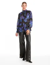 Blusa de gasa plisada con estampado floral en tonos azules y violetas sobre fondo negro. Presenta cuello alto, mangas largas con puños abotonados y un diseño fluido con textura plisada.
