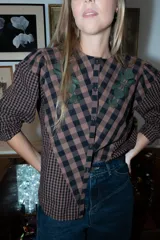 Blusa de algodón con estampado a cuadros en tonos azul y negro, con bordados decorativos en color bordó sobre el canesú. Presenta cierre frontal con botones, cuello redondo y mangas largas levemente abullonadas.