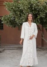 Vestido largo color crudo, estilo camisero, con cuello en V y volados verticales en el frente. Tiene mangas largas abullonadas y corte amplio.