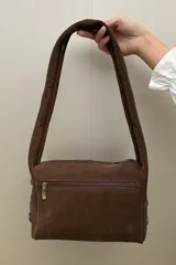 Cartera de hombro confeccionada en gamuza color habano, con diseño de correa ancha decorada con tachas metálicas circulares. Presenta cierre superior y placa metálica con logo en el frente.