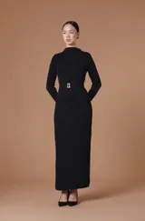 Vestido largo entallado de color negro, con cuello alto y mangas largas. Presenta una textura plisada con ondas verticales y un cinturón integrado con hebilla metálica dorada en la cintura.