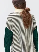 Pullover de tejido acanalado grueso con cuello alto y cierre frontal parcial. Presenta un diseño de bloques de color, con cuerpo en tono beige y mangas en contraste con rayas verticales en tonos rojizos. Incluye un detalle bordado en el pecho.