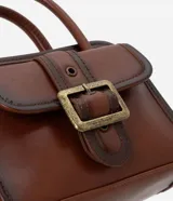 Cartera pequeña de mano con diseño estructurado, confeccionada en poliuretano color marrón con acabado envejecido. Cuenta con tapa frontal decorada con una hebilla metálica, cierre principal con cremallera, asa de mano rígida y correa larga ajustable y desmontable.