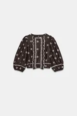 Blusa marrón oscuro con estampado de lunares blancos, manga abullonada por debajo del codo y detalles de bordados perforados y volantes en la parte delantera. Presenta cuello redondo y acabado elástico.