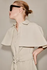 Vestido camisero midi de algodón con diseño de capa, cuello solapa, manga corta tipo capa y trabillas en los hombros. Presenta cierre frontal con botones, bolsillos delanteros y cinturón ajustable del mismo tejido con lazada. El bajo tiene un acabado en línea evasé.