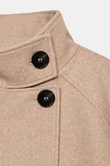 Chaqueta corta de paño en color beige, con cuello subido, manga larga y cierre frontal cruzado con botones oscuros. Presenta bolsillos delanteros de vivo.