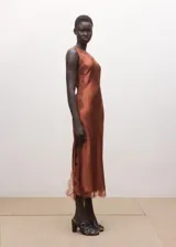 Vestido midi asimétrico de tejido satinado en color tostado, con un tirante fino y otro hombro descubierto. Cuenta con delicados ribetes de encaje a tono en el escote y en el bajo, y una sutil abertura lateral.