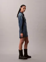 Vestido mini de denim azul con efecto desgastado, de manga larga, con cuello camisero, cierre frontal de botones metálicos y bolsillos con solapa en el pecho.