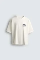 Camiseta de manga corta y cuello redondo, color blanco hueso, con un pequeño parche estampado en el pecho con texto en azul oscuro.