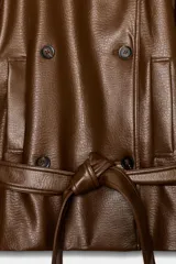 Trench corto de efecto piel color marrón, con cuello de solapa, cierre frontal cruzado con botones y cinturón lazada del mismo material. Presenta mangas largas con trabillas en hombros y puños, y bolsillos de vivo en el delantero.
