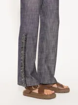 Pantalón de lino con apariencia denim, tiro bajo y corte recto. Presenta detalles de galón con flecos y cadenas en los laterales inferiores.