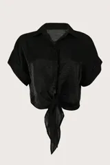Camisa crop negra de tejido fluido y acabado brillante, con cuello camisero, mangas cortas con vuelta y nudo frontal en el dobladillo.