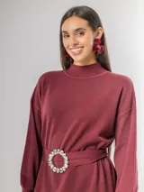 Vestido tipo polerón de tejido de punto color borgoña, con cuello alto, mangas largas con puños ajustados y corte recto. Incluye un cinturón del mismo material con una hebilla decorativa de flor con cristales.