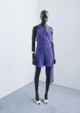 Vestido corto de estilo cruzado con estampado de lunares, cuello en pico y diseño sin mangas. Presenta un lazo lateral para ajuste y falda con caída fluida.