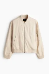 Campera bomber en mezcla de algodón y nylon color beige, con cuello elevado acanalado y cierre frontal de cremallera. Cuenta con bolsillos laterales y un bolsillo con cierre en la manga izquierda. Puños y bajo en punto acanalado, de corte holgado.