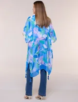 Kimono de gasa azul con estampado botánico de hojas tropicales en tonos celestes, verdes y rosados. Presenta un diseño abierto al frente con mangas cortas y amplias, ruedo asimétrico estilo pañuelo y detalle de borlas en las esquinas inferiores.