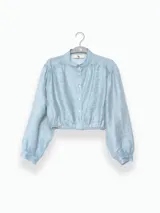 Blusa corta de tejido arrugado liviano, con cuello camisero, cierre frontal de botones y mangas largas abullonadas con puños. Presenta un ruedo elastizado que le otorga una silueta amplia y relajada.