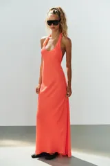 Vestido largo color salmón con cuello halter, escote redondo y espalda descubierta. Cuenta con bajo acabado asimétrico, cierre en el cuello con botón y cremallera oculta en la espalda.