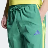 Pantalón deportivo Adidas de corte estándar, color verde con las icónicas tres rayas laterales en color amarillo. Presenta logo de la marca bordado en violeta y bolsillos laterales.