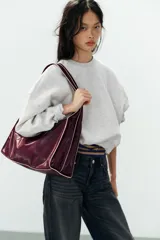 Bolso tipo shopper en formato horizontal, de efecto acharolado en color bordó con detalles de pespuntes en contraste. Cuenta con asa ancha de hombro, bolsillo interior con cierre y cierre principal con botón imantado.