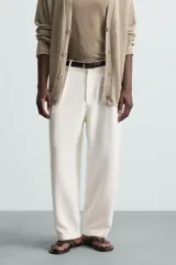 Pantalón blanco de corte relajado (relaxed fit) con pliegues frontales, bolsillos laterales y bolsillos traseros de vivo. Cierre frontal con cremallera y botón.