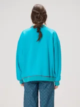 Buzo oversize color gris melange con corte crew, cuello redondo, mangas largas y detalles bordados en color verde en el cuello, puños y ruedo. Presenta el logo de la marca bordado en el pecho.