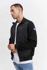 Campera de estilo deportivo con cierre frontal, confeccionada en una combinación de nylon y algodón. Presenta un diseño con paneles texturizados en las mangas, cuello alto y un parche decorativo con coordenadas en el pecho.