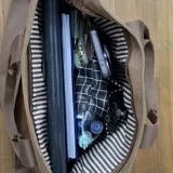 Bolso tipo tote bag de lona color negro, con múltiples bolsillos exteriores y asas cortas y correa larga de tela color marrón. Presenta un parche de cuero marrón con logo grabado en el frente.