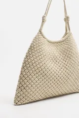 Bolso tipo shopper color crudo, confeccionado en tejido trenzado de algodón con diseño de macramé. Cuenta con dos asas de hombro ajustables que funcionan como cierre del compartimento principal.