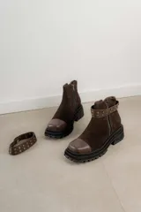 Bota corta de gamuza marrón con puntera de cuero liso, suela track negra y correa desmontable con tachas metálicas.