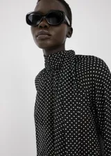 Blusa de poliéster color negro con estampado de lunares blancos, cuello alto con detalle de lazo, manga larga con puños elásticos y diseño recto.