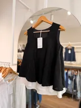 Blusa sin mangas de doble capa, con la capa superior negra de textura sutil y corte evasé, y una capa inferior blanca plisada que sobresale en el ruedo. Presenta escote redondo y diseño amplio.