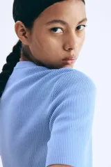 Cárdigan corto de punto acanalado en color celeste, con cuello redondo, manga corta y cierre frontal de botones dorados. Presenta dos bolsillos delanteros decorativos con botón dorado.