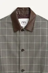 Trench de corte relajado con estampado de cuadros en tonos beige y marrón, cuello solapa en piel marrón, manga larga, bolsillos de vivo en cadera, cinturón ajustable y cierre frontal con botones.