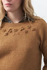 Sweater de tejido suave color tostado, con cuello redondo y detalle de flores bordadas en relieve en la parte superior del pecho. Presenta un corte relajado y mangas largas con puños ajustados.
